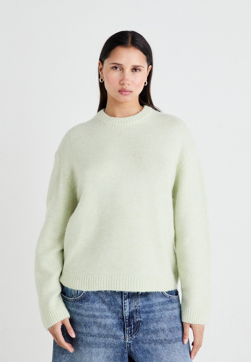 Lys grønn strikket genser med ribbestrikket hals og ermer, med en avslappet passform og myk, teksturert stoff. Brukt med blå jeans.