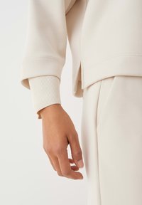 Lichtbeige sweatshirt met geribbelde manchetten en zijsplitten, gemaakt van gladde stof en met een losse pasvorm. Close-up van een hand op de stof.