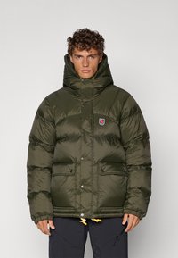 Veste doudoune verte avec un col haut, fermeture éclair et pression, deux poches avant et un patch logo. Tissu texturé et brillant avec un ourlet ajustable.