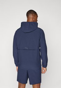 Marineblå aktivjakke med hette, elastiske mansjetter og bakventil. Materialet ser lett og pustende ut. Kombinert med matchende shorts.