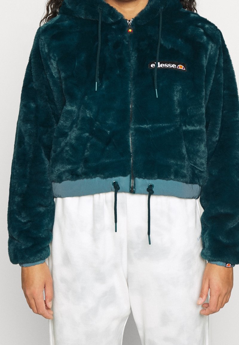 Turkusowa pluszowa kurtka typu zip-up o skróconym kroju, z naszywką z logo, sznurkiem w talii oraz miękką, teksturowaną powierzchnią.