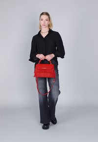 Borsa a tracolla rossa in pelle con dettagli a frange, forma rettangolare, manico superiore e una tracolla staccabile, abbinata a una blusa nera e jeans.