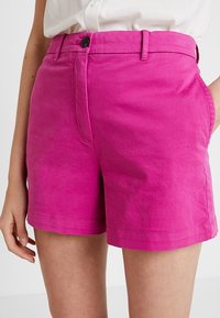 Pantalones cortos de algodón rosa con una cintura ajustada, cierre de botón y bolsillos laterales. La tela tiene una textura suave y detalles de costura mínimos.