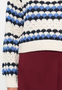 Strickpullover in Creme mit navy- und blauen Zickzackmustern, mit einem gerippten Saum. Kombiniert mit einer weinroten Unterhose.