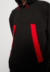 Felpa nera con cappuccio, caratterizzata da due strisce rosse verticali con il logo "HUGO". Il materiale sembra morbido, con una finitura testurizzata e cappuccio con coulisse regolabili.