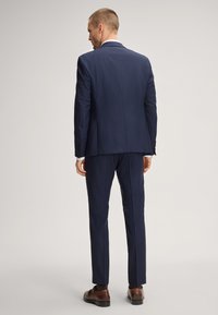 Completo blu navy con giacca sartoriale a singolo spacco e pantaloni abbinati. Tessuto liscio con una lieve lucentezza, indossato con scarpe marroni.