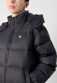 Svart dunjakke med høy krage, glidelås og dekorativ knappdetalj på den ene skulderen. Har en liten logo på brystet.