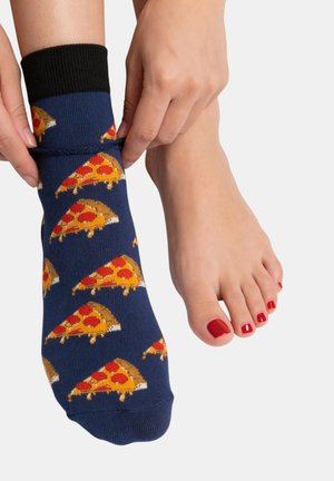 Des mains tirant sur une chaussette bleu marine ornée de motifs de tranches de pizza, à côté d’un pied nu avec des ongles d’orteils peints en rouge sur un fond uni.