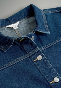 Denima krekls tumši zilā krāsā, ar metāla pogām un norādītu apkakli. Raksturīgas kontrastējošas šuves un regulējama apkakles aizdare.