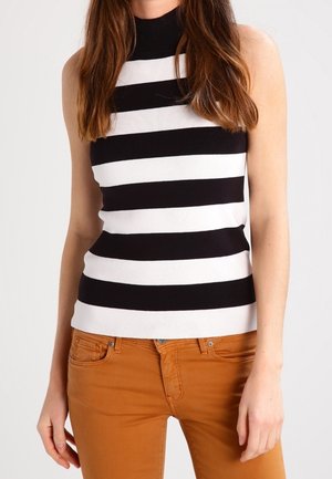 Kvinde iført en ærmeløs top med sorte og hvide horisontale striber og sennepsfarvede bukser mod en ensfarvet baggrund.