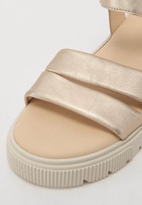 Sandaler med metallisk gyllen syntetisk overdel, som har to brede stropper og en tan fotseng. Teksturert hvit gummisåle med riller.