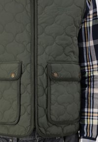 Zaļa izšūta veste ar divām pogām paredzētām priekšējām kabatām, ar teksturētu rakstu un logotipu uz krūtīm. Valkāta virs raibiem krekliem.