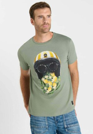 Man draagt lichtgroen T-shirt met afbeelding van een geel gestreepte helm, zonnebril en baard gemaakt van citroenen en witte bloemen.