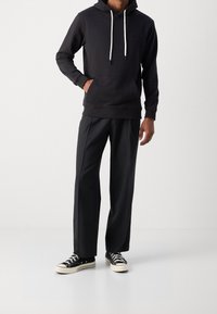 Sudadera negra con bolsillo delantero y cordones blancos, combinada con pantalones oscuros ajustados y zapatillas negras con suelas blancas.