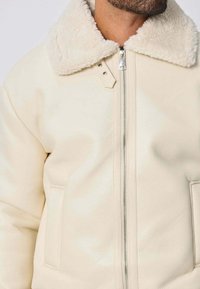 Veste en cuir couleur crème avec un col en fausse fourrure, dotée d'une fermeture éclair sur le devant et de deux poches latérales. Texture lisse et design minimaliste.