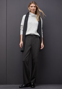 Suéter gris de punto con un cuello ancho, combinado con pantalones oscuros de rayas. Una bufanda oscura cuelga de manera suelta, y unos zapatos negros completan el conjunto.