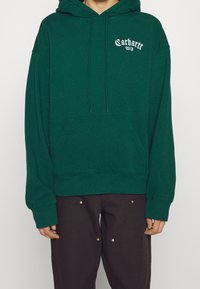 Sudadera con capucha verde hecha de algodón suave; cuenta con un bolsillo frontal, capucha con cordón y el logo "Carhartt WIP" en blanco. Corte holgado con puños acanalados.