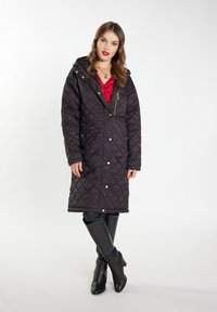 Cappotto trapuntato nero con dettagli in oro, cappuccio, maniche lunghe e chiusure a pressione. Indossato sopra una maglia rossa e abbinato a stivali neri fino al ginocchio.