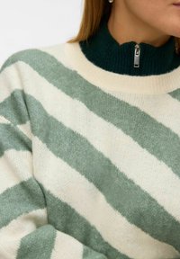 Sweter w paski wykonany z miękkiego, dzianinowego materiału w zielono-kremowe ukośne paski, z wysokim ściągaczem przy szyi i zapięciem na zamek.