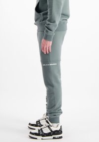 Sweatshirt et joggers verts, en tissu doux. Les joggers disposent de poches latérales et du logo "BLCK BNNS". Les baskets visibles sont noires avec des accents blancs.