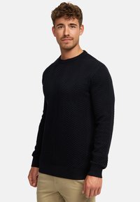 Marineblauwe gebreide trui met een gestructureerd visgraatpatroon, ronde hals en geribbelde manchetten. Gedragen over lichtgekleurde broek.