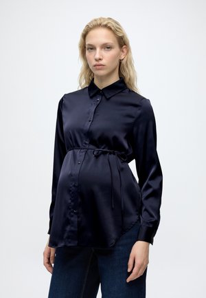 Jeune femme enceinte aux longs cheveux blonds, portant un chemisier de maternité en satin bleu marine à boutons et un jean foncé, debout sur fond blanc.
