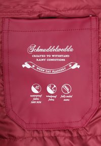 Czerwana wodoodporna etykieta z brandingiem 'Schmuddelwedda', informująca o 3000 mm odporności na wodę, wiatroszczelności i w pełni uszczelnionych szwach.