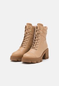 Un par de botas de tobillo en tono marrón claro, con cordones, tacones gruesos, cuellos acolchados y paneles laterales texturizados, sobre un fondo blanco liso.