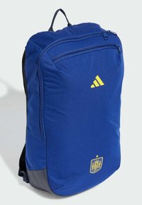 Mochila de poliéster azul con un asa superior, bolsillo frontal con cremallera y logo amarillo de Adidas. Presenta un emblema con un detalle de corona y estrella.