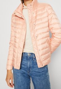 Kurtka pikowana w miękkim odcieniu brzoskwini, z wysokim kołnierzem, zamkiem błyskawicznym i zatrzaskami. Materiał ma błyszczące wykończenie oraz pikowaną fakturę.