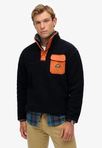 Marineblauer Fleecepullover mit orangefarbenem Kragen und Brusttasche, mit strukturierter Oberfläche und Berg-Logo. Tragend über einem karierten Hemd.