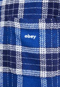 Blå og hvit rutete stoff med en strukturert overflate. Har en liten hvit "obey"-logo, tydelig plassert på materialet.