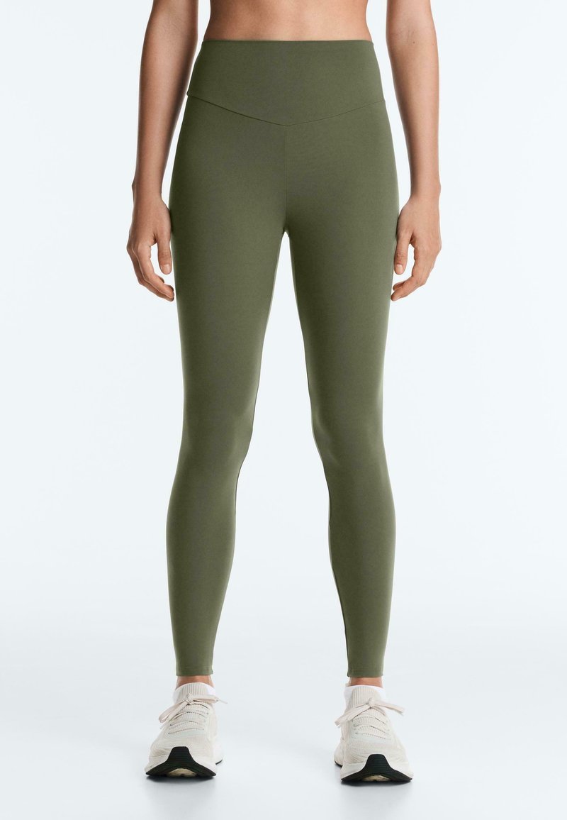 Femme portant des leggings taille haute olive et des baskets blanches, debout, les bras détendus le long du corps, sur un fond uni.