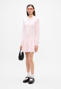 Polo šaty v jemnej ružovej látke s dlhými rukávmi a plisovanou sukňou. Doplnky zahŕňajú čiernu kabelku a robustné čierne topánky s bielymi ponožkami.
