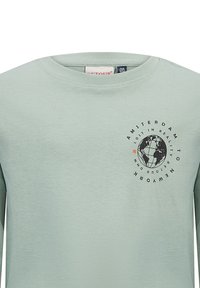 Helles grünes Langarm-T-Shirt mit Rundhalsausschnitt. Es hat ein schwarzes Globus-Graphic und den Text "AMSTERDAM NACH NEW YORK" im kreisförmigen Format.
