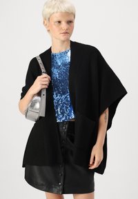 Svart stickad cardigan med breda ärmar, som bärs över en blå paljett-topp. Har en avslappnad passform. Modellen håller en liten metallisk silverväska.