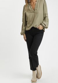 Olivengrünes, langärmliges Blusenshirt mit V-Ausschnitt und gerafften Bündchen, kombiniert mit hochgeschnittenen schwarzen Jeans und beigen Stiefeletten.