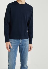 Granatowy sweter z długim rękawem wykonany z miękkiej tkaniny, z okrągłym dekoltem i luźnym krojem, zestawiony z jasnoniebieskimi jeansami. Dyskretne logo na piersi.