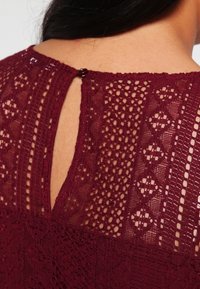 Burgundy topp med spets bak i geometriska mönster, en nyckelhålöppning och en liten rund knappstängning vid halsringningen.