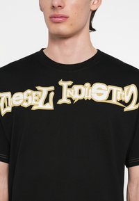 Μαύρο βαμβακερό μπλουζάκι με έντονο, στιλιζαρισμένο λευκό και χρυσό κείμενο "Diesel Industry" στο στήθος. Ομαλή υφή και κοντά μανίκια.