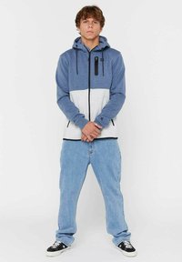 Felpa con cappuccio in design a blocchi di colore blu e grigio, chiusura con zip, lacci regolabili e tasca con zip. I jeans sono di un blu chiaro.
