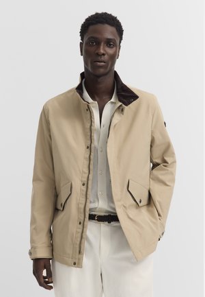 Mann trägt beige Jacke über weißem Hemd und Hose, steht vor einem einfarbigen hellen Hintergrund mit einer Hand in der Tasche.