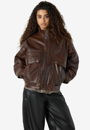 Femme aux longs cheveux bouclés portant une veste en cuir marron zippée et un pantalon en cuir noir, debout devant un fond uni.