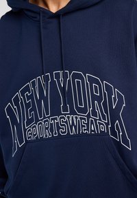 Sweat à capuche bleu marine avec texte imprimé blanc indiquant « NEW YORK SPORTSWEAR ». Comprend une poche kangourou et une capuche avec cordon de serrage.