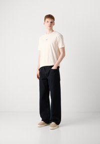 Kremfarget kortermet t-skjorte med lite logo, kombinert med løstsittende mørk marineblå bukser og lysebeige joggesko. Glatt stofftekstur.