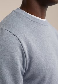 Sudadera de cuello redondo en gris claro con una tela suave y texturizada, que presenta puños y cuello acanalados. Vista en primer plano de los detalles de la costura del hombro.