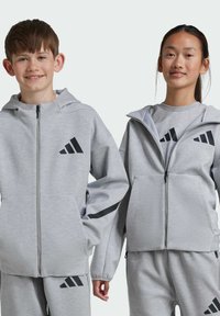 Dos niños llevan chaquetas con capucha de cremallera en gris claro a juego, con detalles en negro y el logo de Adidas. Las chaquetas tienen bolsillos laterales y puños acanalados.