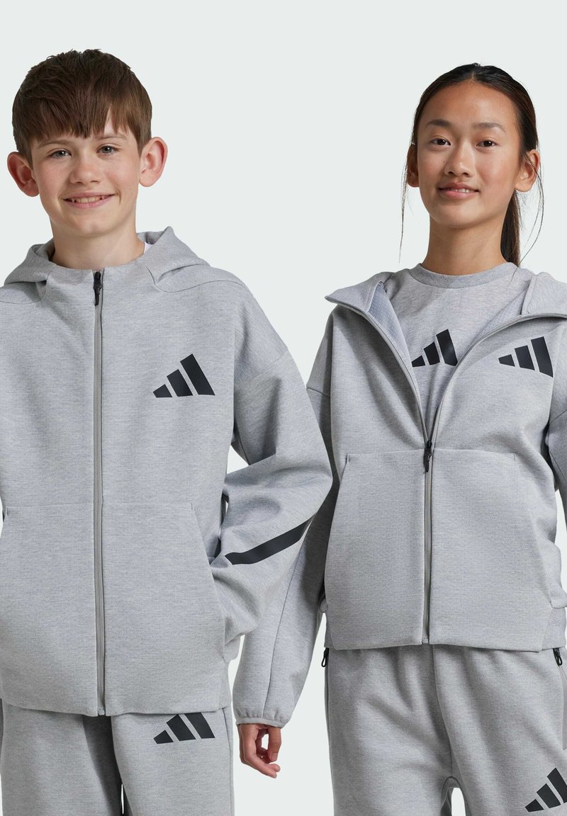 Dos niños llevan chaquetas con capucha de cremallera en gris claro a juego, con detalles en negro y el logo de Adidas. Las chaquetas tienen bolsillos laterales y puños acanalados.