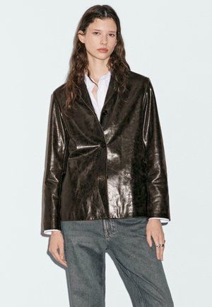 Blazer en cuir marron avec une finition brillante, doté d'une fermeture à un bouton et de deux poches, porté sur une chemise blanche et un jean gris.