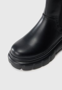 Melnas ādas zābaks ar masīvu gumijas zoli un elastīgu sānu paneli, rādīts tuvu plānā uz baltā fona.